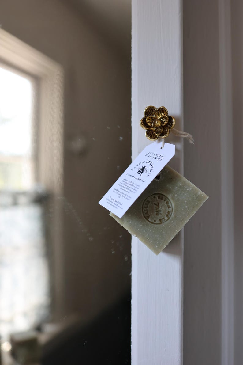 Puede incluir: Un pomo dorado en forma de flor sujeta una etiqueta y una pastilla de jab&oacute;n verde. La etiqueta dice "Lavender & Linen Co." y "Savon de Carmel." El jab&oacute;n tiene un dise&ntilde;o estampado. El fondo es un marco de puerta blanco.