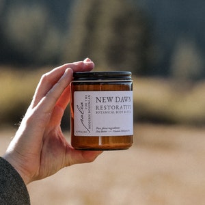 Może przedstawiać: Brązowy szklany słoik z białą etykietą, na kt&oacute;rej widnieje napis "New Dawn Restorative Botanical Body Butter". Etykieta zawiera r&oacute;wnież tekst "For the Modern Bohemian" i "Pure plant ingredients Shea Butter - Vitamin E & Jojoba Oil".