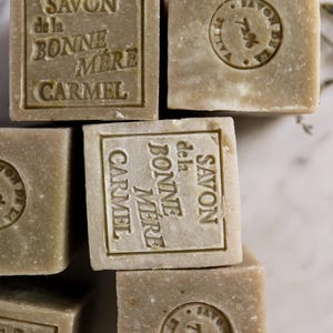 Puede incluir: Primer plano de varios jabones cuadrados de color verde oliva. Cada jab&oacute;n est&aacute; estampado con las palabras "SAVON de la BONNE MERE CARMEL" en un marco rectangular. Algunos jabones tienen un sello circular con las palabras "SAVON DE LA VALLEE 72%."