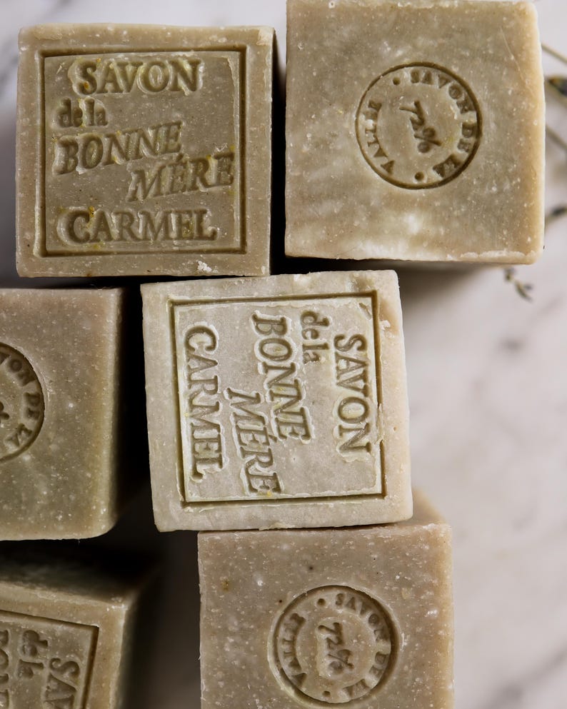Puede incluir: Primer plano de varios jabones cuadrados de color verde oliva. Cada jab&oacute;n est&aacute; estampado con las palabras "SAVON de la BONNE MERE CARMEL" en un marco rectangular. Algunos jabones tienen un sello circular con las palabras "SAVON DE LA VALLEE 72%."