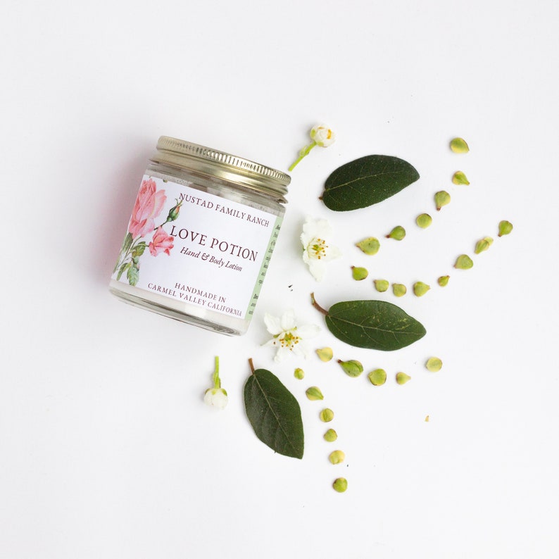 May include: A white jar with a gold lid and a label that reads "NUSTAD FAMILY RANCH LOVE POTION Hand & Body Lotion HANDMADE IN CARMEL VALLEY CALIFORNIA". The jar is surrounded by white flowers and green leaves on a white background.