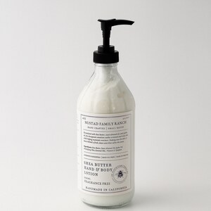 May include: A clear glass bottle with a black pump dispenser filled with white lotion. The bottle has a white label with black text that reads "NUSTAD FAMILY RANCH HAND CRAFTED | SMALL BATCH SHEA BUTTER HAND & BODY LOTION SCENT: FRAGRANCE FREE HANDMADE IN CALIFORNIA".