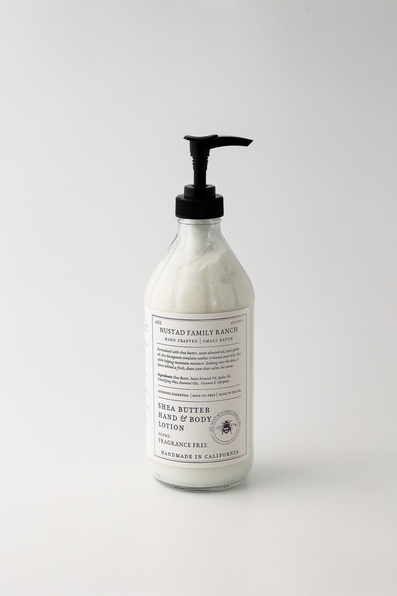 May include: A clear glass bottle with a black pump dispenser filled with white lotion. The bottle has a white label with black text that reads "NUSTAD FAMILY RANCH HAND CRAFTED | SMALL BATCH SHEA BUTTER HAND & BODY LOTION SCENT: FRAGRANCE FREE HANDMADE IN CALIFORNIA".
