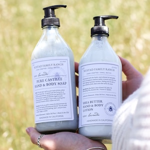 May include: Two bottles of lavender-scented body care products from Nustad Family Ranch. The bottle on the left is a clear glass bottle with a pump dispenser and a label that reads "Pure Castile Hand & Body Soap". The bottle on the right is a clear glass bottle with a pump dispenser and a label that reads "Shea Butter Hand & Body Lotion". Both bottles are labeled "Handmade in California".