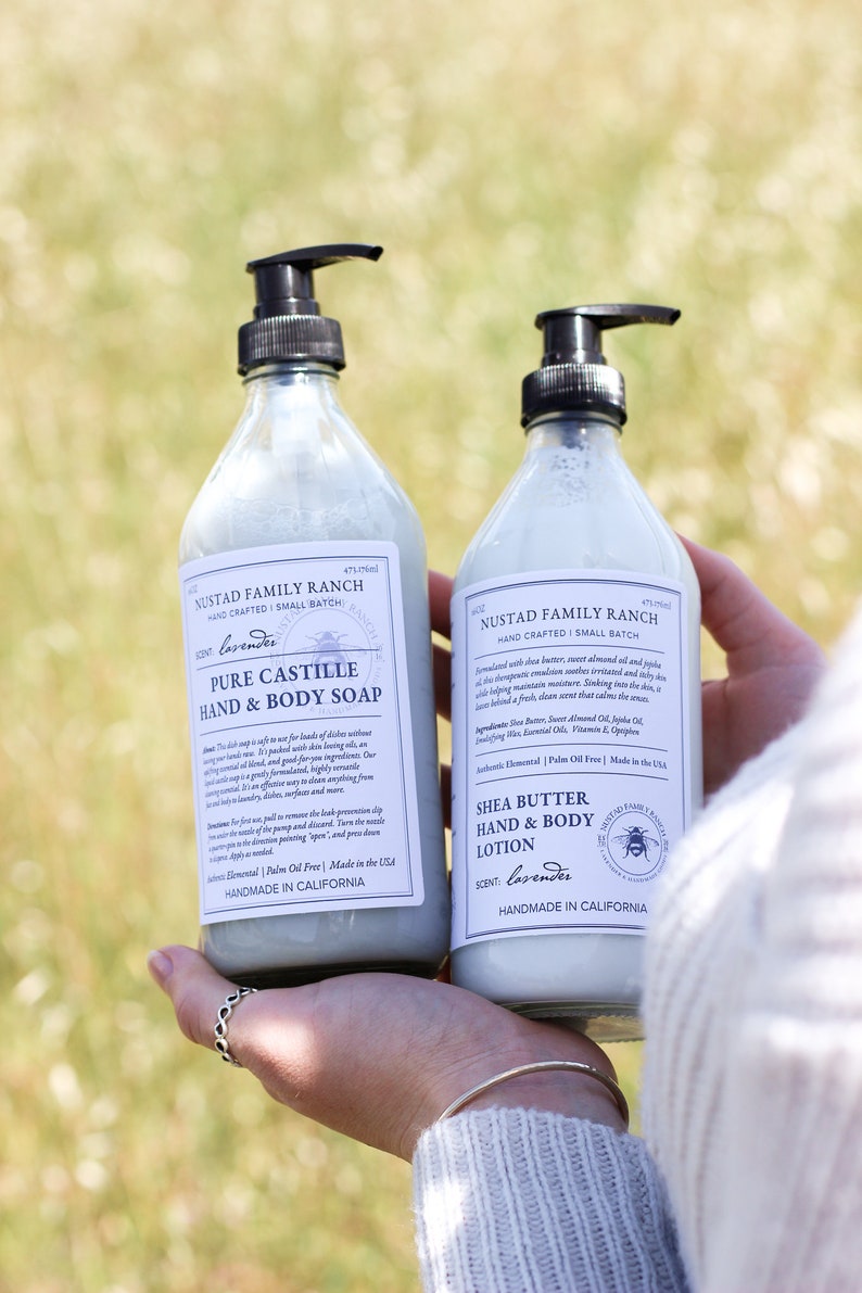 May include: Two bottles of lavender-scented body care products from Nustad Family Ranch. The bottle on the left is a clear glass bottle with a pump dispenser and a label that reads "Pure Castile Hand & Body Soap". The bottle on the right is a clear glass bottle with a pump dispenser and a label that reads "Shea Butter Hand & Body Lotion". Both bottles are labeled "Handmade in California".