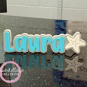 May include: A custom-made name sign that reads "Laura" in a playful, rounded font. The letters are a vibrant turquoise, outlined in a soft gray, and accompanied by a white starfish. The sign is likely a decorative item, possibly for a child's room or a personalized gift.