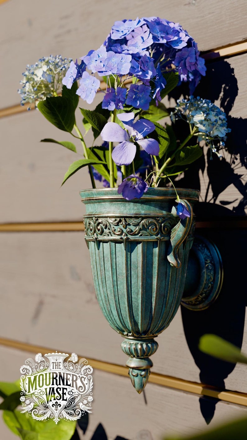 Peut inclure: Un vase mural orn&eacute; de couleur vert sarcelle avec une poign&eacute;e d&eacute;corative. Le vase est rempli d'un bouquet d'hortensias violets et bleus.