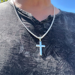 May include: A silver cross pendant necklace with a rope chain. The cross is a simple, polished design. The necklace is worn against a dark gray t-shirt.