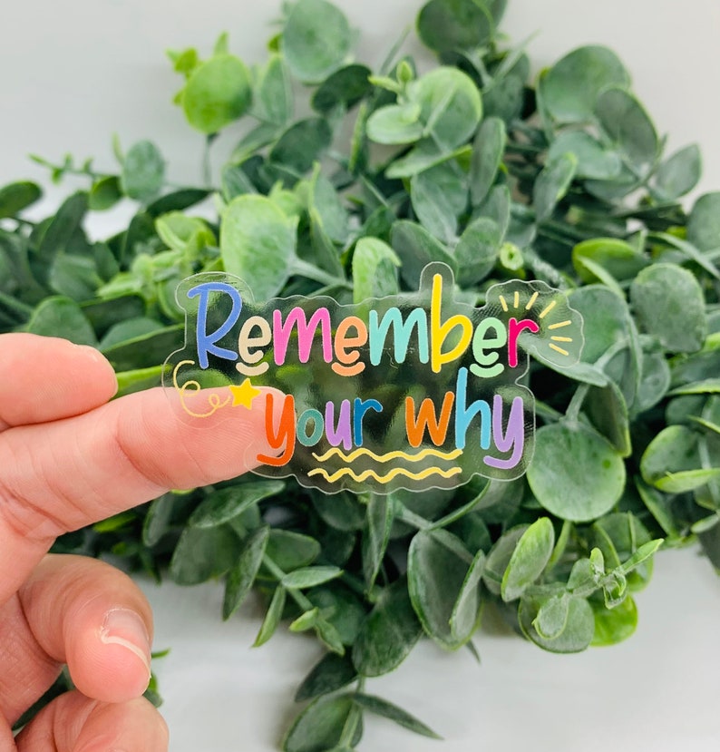 May include: A clear sticker with the text "Remember your why" in colourful lettering. The sticker has a wavy, transparent border and is held against a backdrop of green foliage.