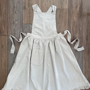 May include: A light beige apron with a pocket and ruffled hem. The apron has a cross-back design and long ties at the waist. The apron is made of a soft, textured fabric and is displayed on a wooden surface.