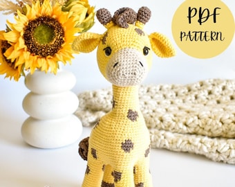 May include: A crocheted giraffe amigurumi toy with brown spots and a brown mane. The giraffe is standing on a white surface with a sunflower bouquet in a white vase in the background. The text "PDF PATTERN" is in a yellow circle in the top right corner of the image.