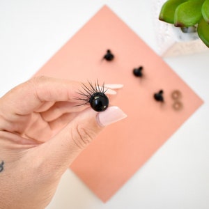 May include: Black plastic safety eyes with black lashes, perfect for crafting plush toys or dolls. The eyes are shown on a pink surface.