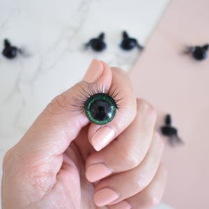 Puede incluir: Primer plano de un ojo de mu&ntilde;eca de pl&aacute;stico verde y negro con pesta&ntilde;as. El ojo est&aacute; siendo sostenido por una mano.