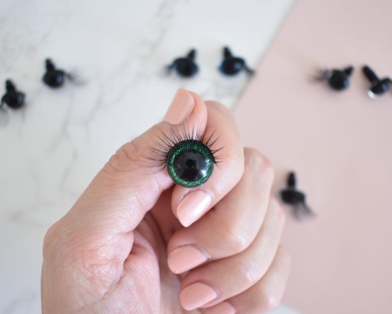 Puede incluir: Primer plano de un ojo de mu&ntilde;eca de pl&aacute;stico verde y negro con pesta&ntilde;as. El ojo est&aacute; siendo sostenido por una mano.