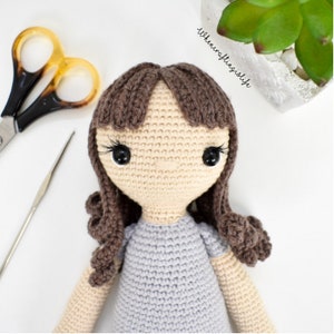 May include: A crocheted doll with brown hair, a light blue dress, and black eyes. The doll is looking directly at the camera.