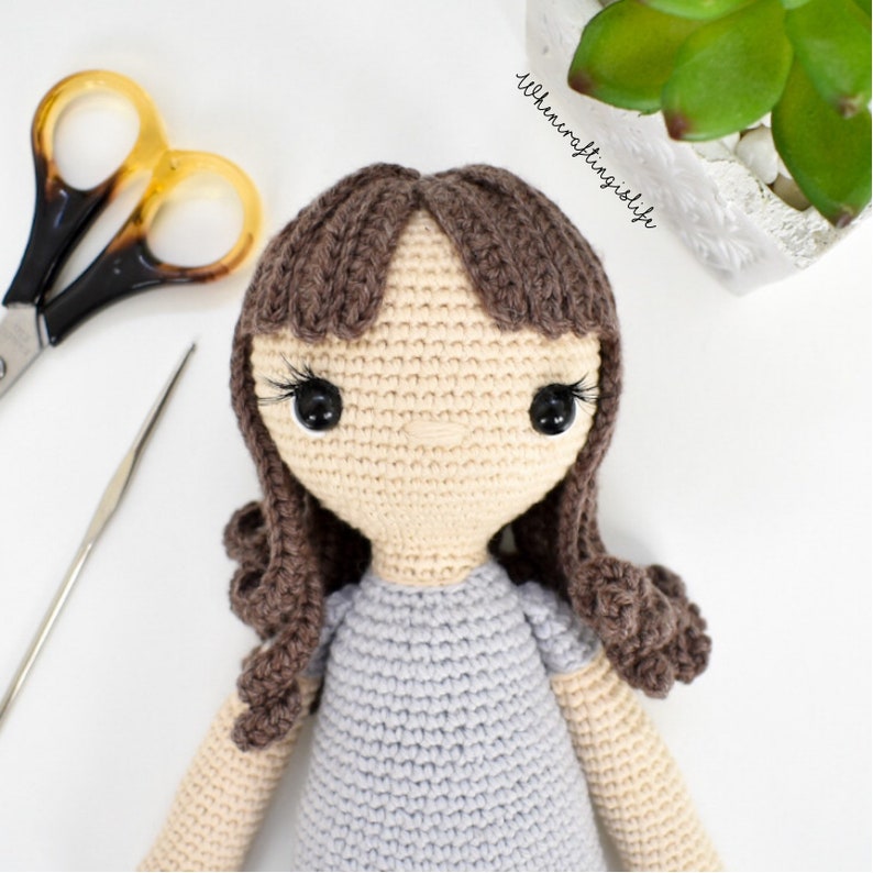 May include: A crocheted doll with brown hair, a light blue dress, and black eyes. The doll is looking directly at the camera.