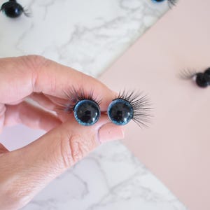 Puede incluir: Dos ojos de mu&ntilde;eca de vidrio azul con pupilas negras y pesta&ntilde;as pegadas. Los ojos se sostienen en una mano.