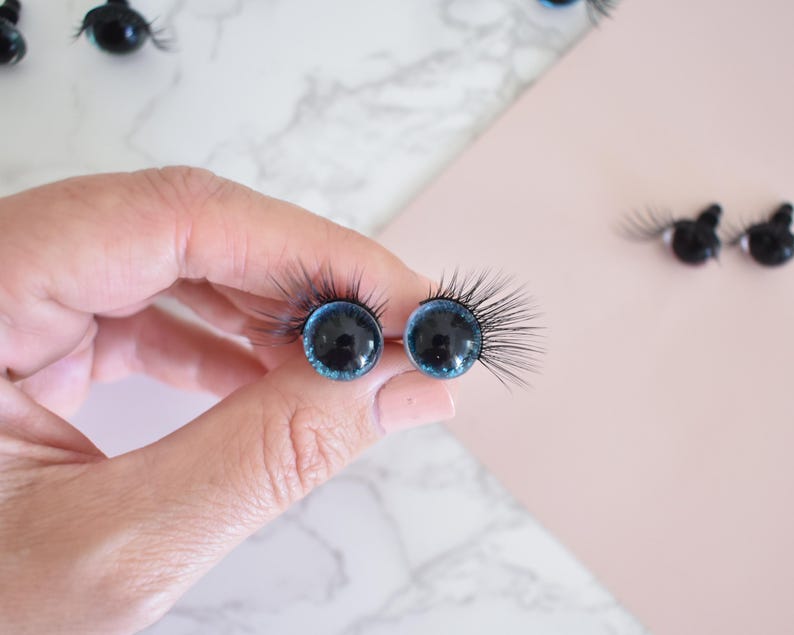 Puede incluir: Dos ojos de mu&ntilde;eca de vidrio azul con pupilas negras y pesta&ntilde;as pegadas. Los ojos se sostienen en una mano.