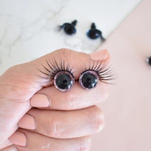 Puede incluir: Dos ojos de muñeca de plástico negro con purpurina rosa alrededor del iris y pestañas negras. Los ojos están siendo sostenidos por una mano.