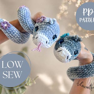 May include: Two plush crochet snakes in shades of blue and grey. One snake is light blue with purple flower accents, the other is grey with a blue accent. The text "PDF PATTERN" and "LOW SEW" are visible.