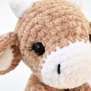 May include: Close-up of a crocheted brown and white cow plush toy with black eyes and eyelashes.