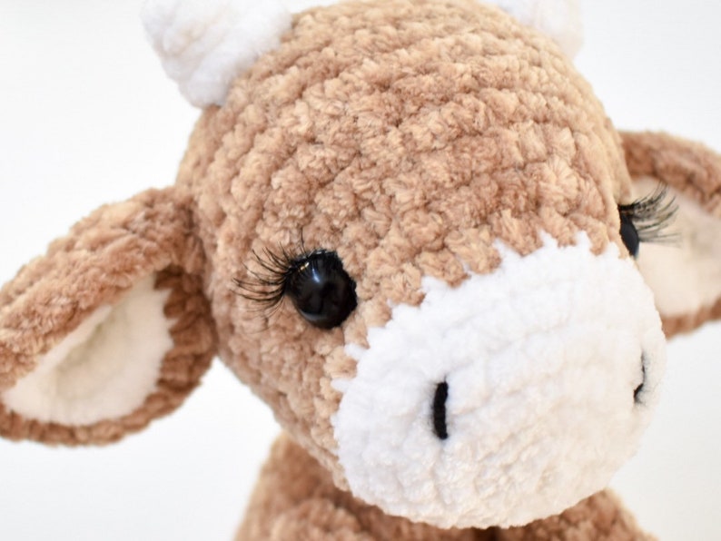May include: Close-up of a crocheted brown and white cow plush toy with black eyes and eyelashes.