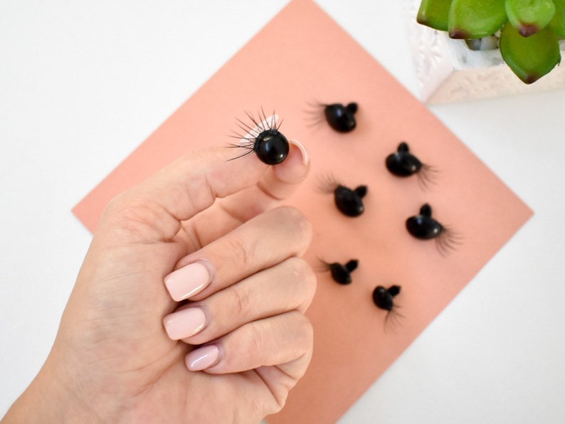 May include: A close-up of black plastic doll eyes with eyelashes. The eyes are on a peach-colored surface.