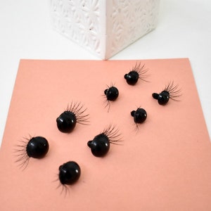 May include: Eight black plastic googly eyes with eyelashes on a peach colored surface.