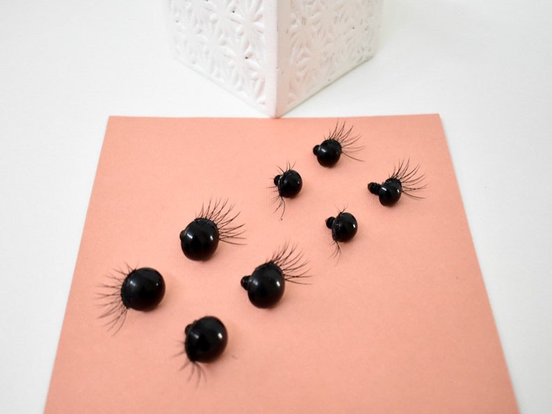 May include: Eight black plastic googly eyes with eyelashes on a peach colored surface.