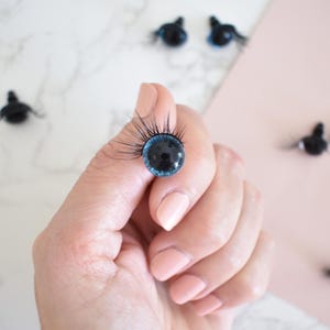 Puede incluir: Primer plano de un ojo de mu&ntilde;eca azul y negro con pesta&ntilde;as. El ojo est&aacute; siendo sostenido por una mano. Hay varios otros ojos de mu&ntilde;eca en el fondo.