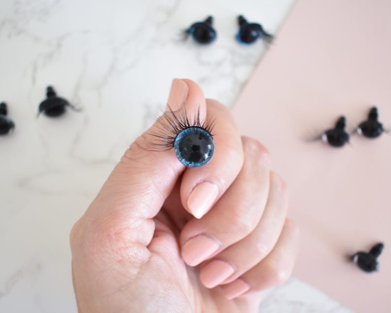 Puede incluir: Primer plano de un ojo de mu&ntilde;eca azul y negro con pesta&ntilde;as. El ojo est&aacute; siendo sostenido por una mano. Hay varios otros ojos de mu&ntilde;eca en el fondo.