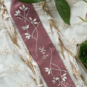 May include: A dusty rose fabric sash with white embroidery of floral sprigs and the word "Mrs." in cursive.