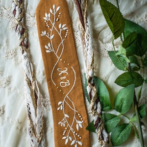 May include: A brown linen bookmark with white embroidery of a floral design and the text "B&B".