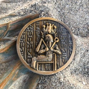 Puede incluir: Una moneda de color bronce que representa a un faraón egipcio sentado en un trono, rodeado de jeroglíficos. La moneda es circular con un diseño en relieve detallado. La moneda descansa sobre una superficie arenosa con un trozo de madera desgastada.