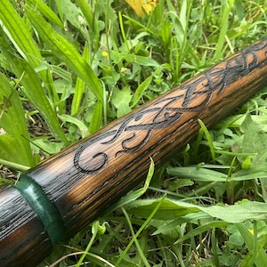 Puede incluir: Un bast&oacute;n de madera con intrincadas tallas de nudos celtas. El bast&oacute;n est&aacute; te&ntilde;ido de un marr&oacute;n c&aacute;lido con detalles oscuros en los detalles tallados. Bandas verdes y marrones acent&uacute;an los extremos, sobre hierba verde.