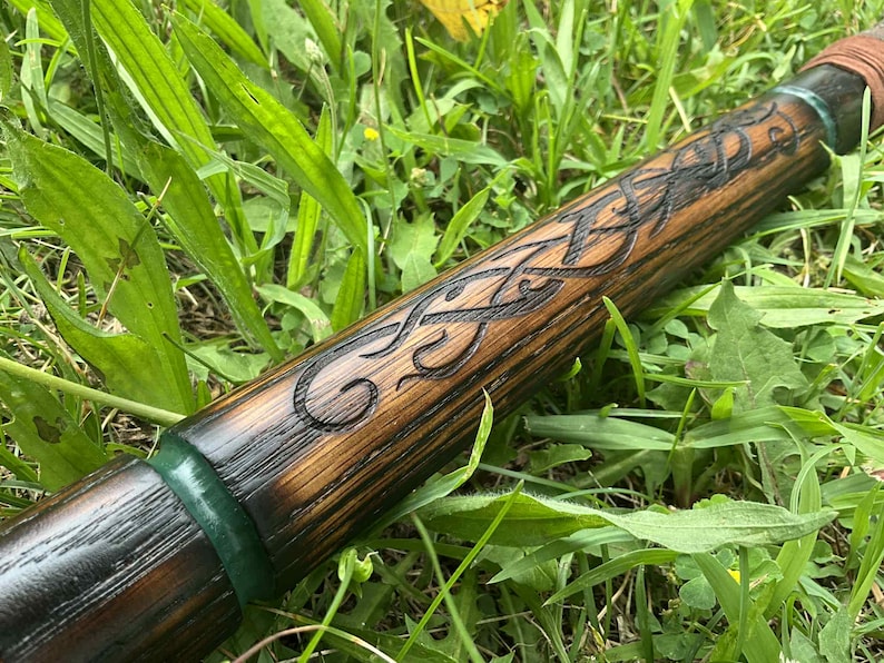 Puede incluir: Un bast&oacute;n de madera con intrincadas tallas de nudos celtas. El bast&oacute;n est&aacute; te&ntilde;ido de un marr&oacute;n c&aacute;lido con detalles oscuros en los detalles tallados. Bandas verdes y marrones acent&uacute;an los extremos, sobre hierba verde.