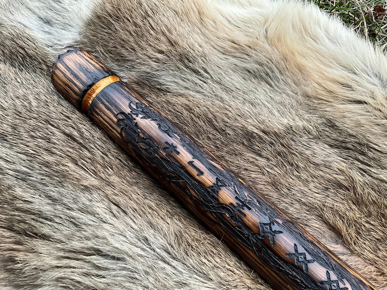 May include: A wooden staff with a dark brown finish and intricate carvings. The staff has a gold band near the top and is decorated with a repeating pattern of geometric shapes.