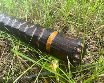 Op de afbeelding: Een handgesneden houten voorwerp met een donkere, verkoold afwerking en een lichtere oranje-bruine band. Het uiteinde heeft verschillende geboorde gaten. Het voorwerp ligt in groen gras.