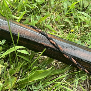 Puede incluir: Un bast&oacute;n de madera con un acabado marr&oacute;n oscuro y una correa trenzada de cuero negro y marr&oacute;n. El bast&oacute;n est&aacute; sobre un lecho de hierba verde.