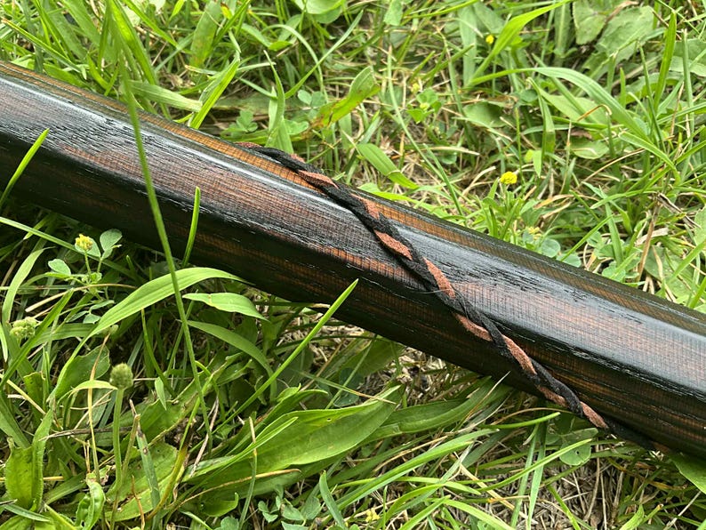 Puede incluir: Un bast&oacute;n de madera con un acabado marr&oacute;n oscuro y una correa trenzada de cuero negro y marr&oacute;n. El bast&oacute;n est&aacute; sobre un lecho de hierba verde.