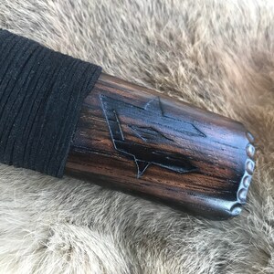 Op de afbeelding: Close-up van een donkerhouten handvat met een zwarte leren wikkeling. Het handvat heeft een gegraveerd ontwerp van een gestileerde pijl met een ster aan de punt.