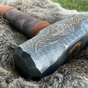 May include: A silver-colored metal hammer with a brown leather handle. The hammer head has an intricate design of a wolf's head and Celtic knotwork.