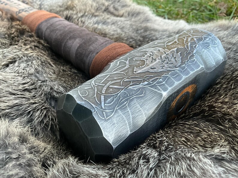 May include: A silver-colored metal hammer with a brown leather handle. The hammer head has an intricate design of a wolf's head and Celtic knotwork.
