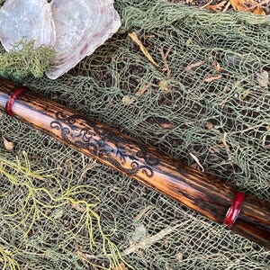 May include: A handcrafted wooden flute with a dark, textured finish and intricate etched designs. The flute features red bands near the ends and rests on a backdrop of fishing net, shells, and greenery.