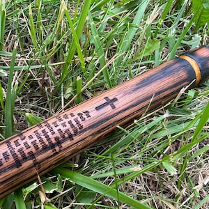 Op de afbeelding: Een cilindervormige houten staf met een donkere, geblakerde afwerking en een natuurlijke houtnerf. De staf is voorzien van een gegraveerd christelijk kruis en de tekst van Johannes 3:16: For God so loved the world that he gave his only begotten Son.