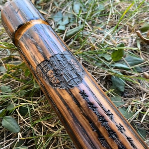 May include: A wooden walking stick with a dark brown stain and a gold band. The stick has a carved design and runes.