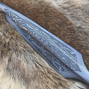 May include: Close-up of a spearhead with detailed etched patterns. The metal blade has intricate designs and a bronze accent. The spearhead is set against a backdrop of brown and tan fur.