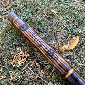 Op de afbeelding: Een houten wandelstok met een donkerbruine vlek en gouden accenten. De stok heeft een gesneden ontwerp en een inscriptie.