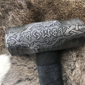 Op de afbeelding: Een zwarte metalen hamer met een decoratief ontwerp en runen op de kop. De steel is omwikkeld met zwart leer.