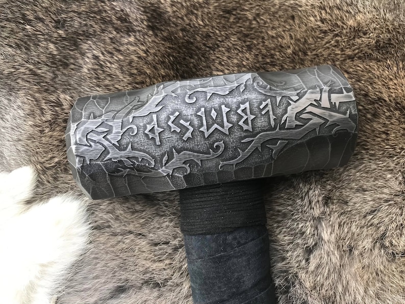 Op de afbeelding: Een zwarte metalen hamer met een decoratief ontwerp en runen op de kop. De steel is omwikkeld met zwart leer.